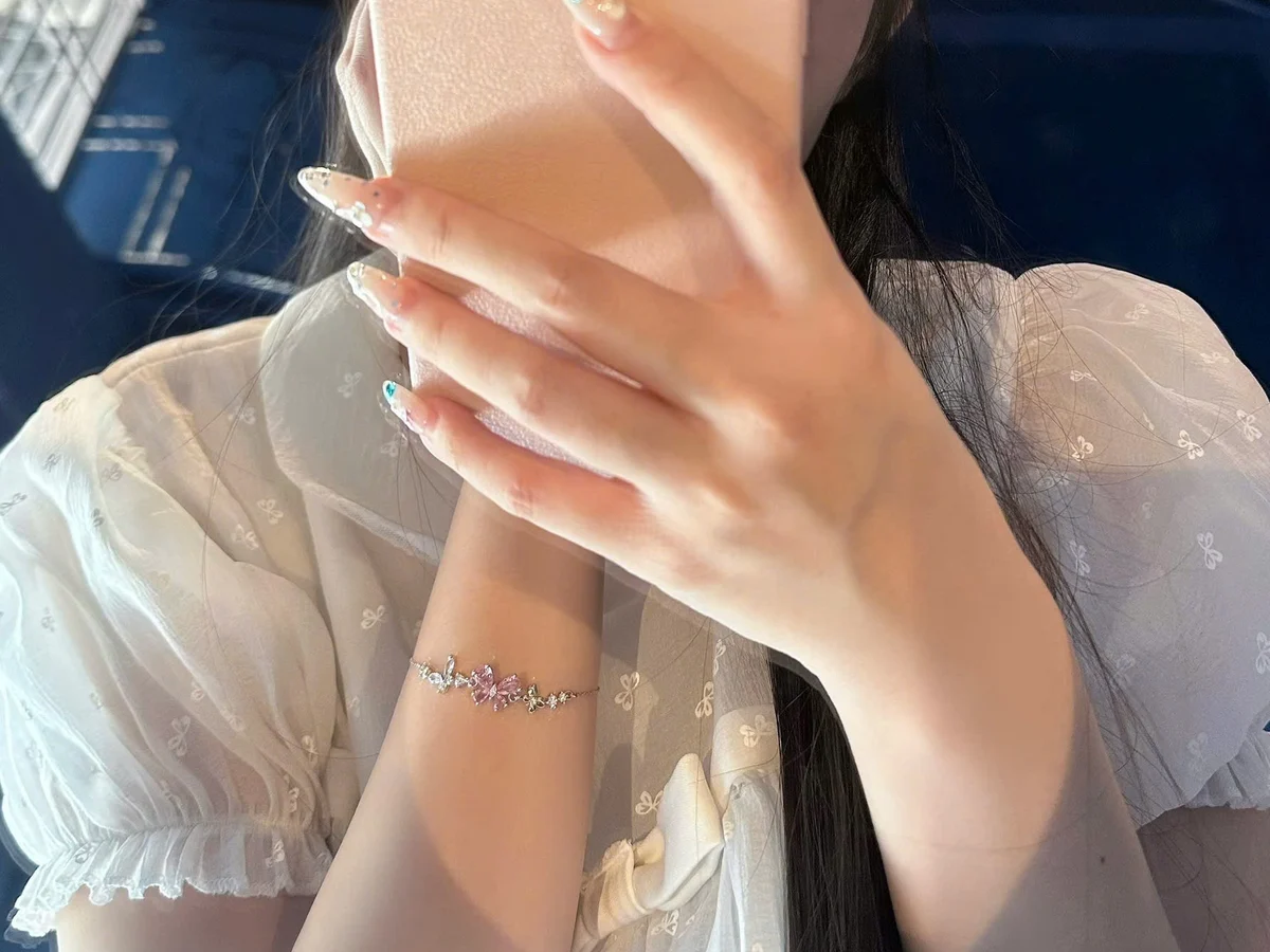 Cổ tay cô gái đeo lắc tay bạc bản mảnh gắn đá hình bướm hồng, tay áo voan trắng — kiểu quà tặng bạn gái cung Xử Nữ đeo được mỗi ngày